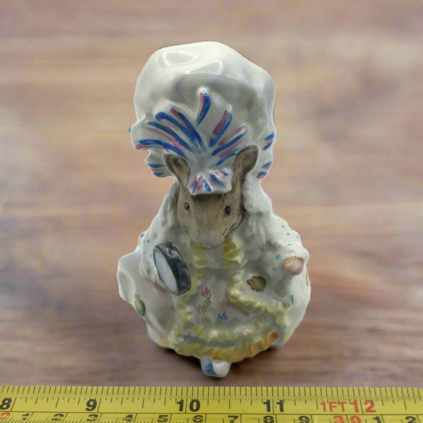 Small ceramic figure of a mouse with a headdress on a wooden surface with a ruler for scale.