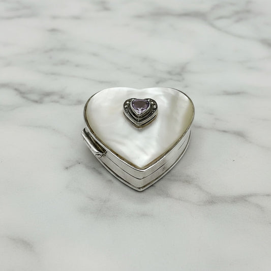 A heart-shaped silver pill box with a white mother-of-pearl exterior and a small heart-shaped purple gemstone in the center, resting on a white marble surface.