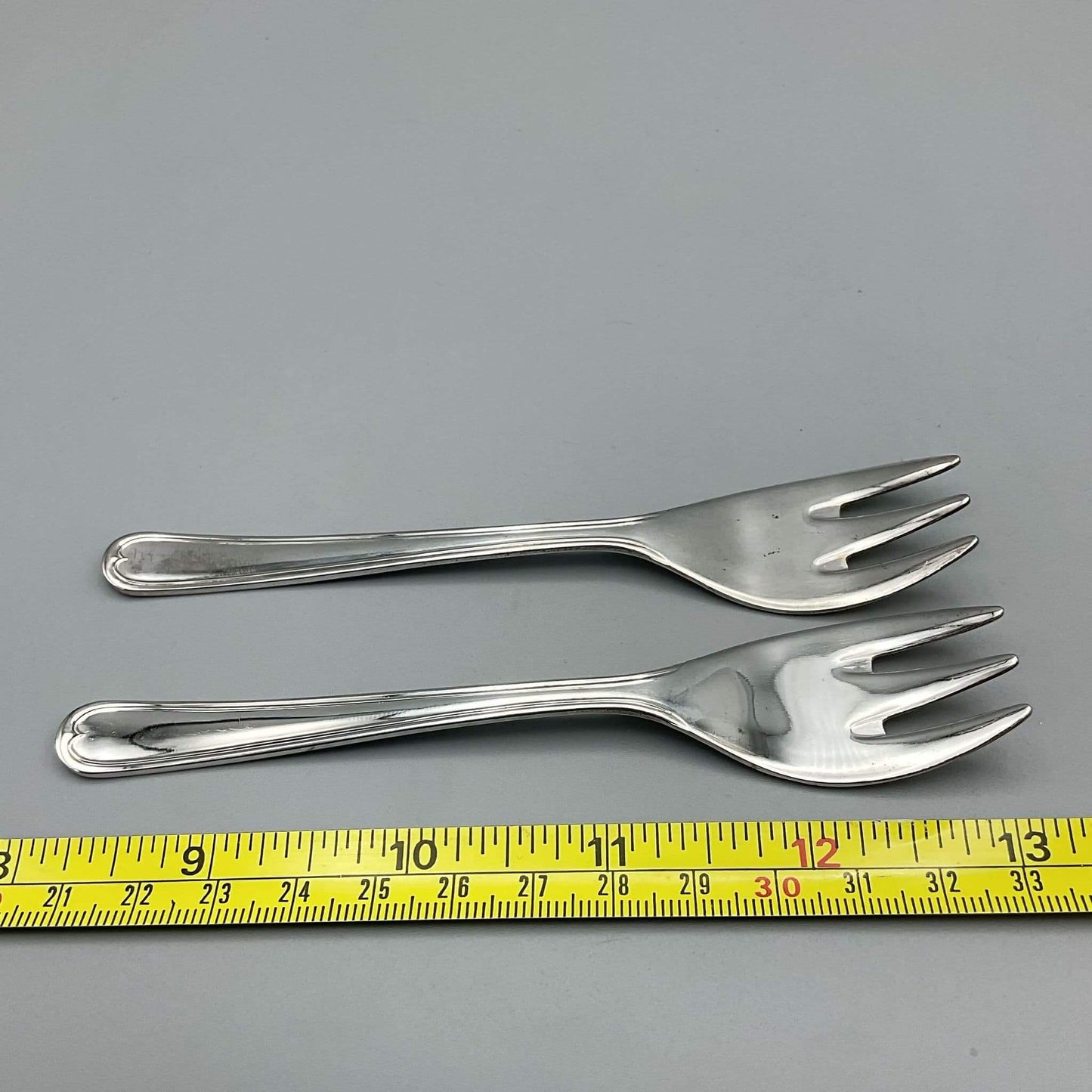 Two silver forks on a gray background with a yellow measuring tape at the bottom.