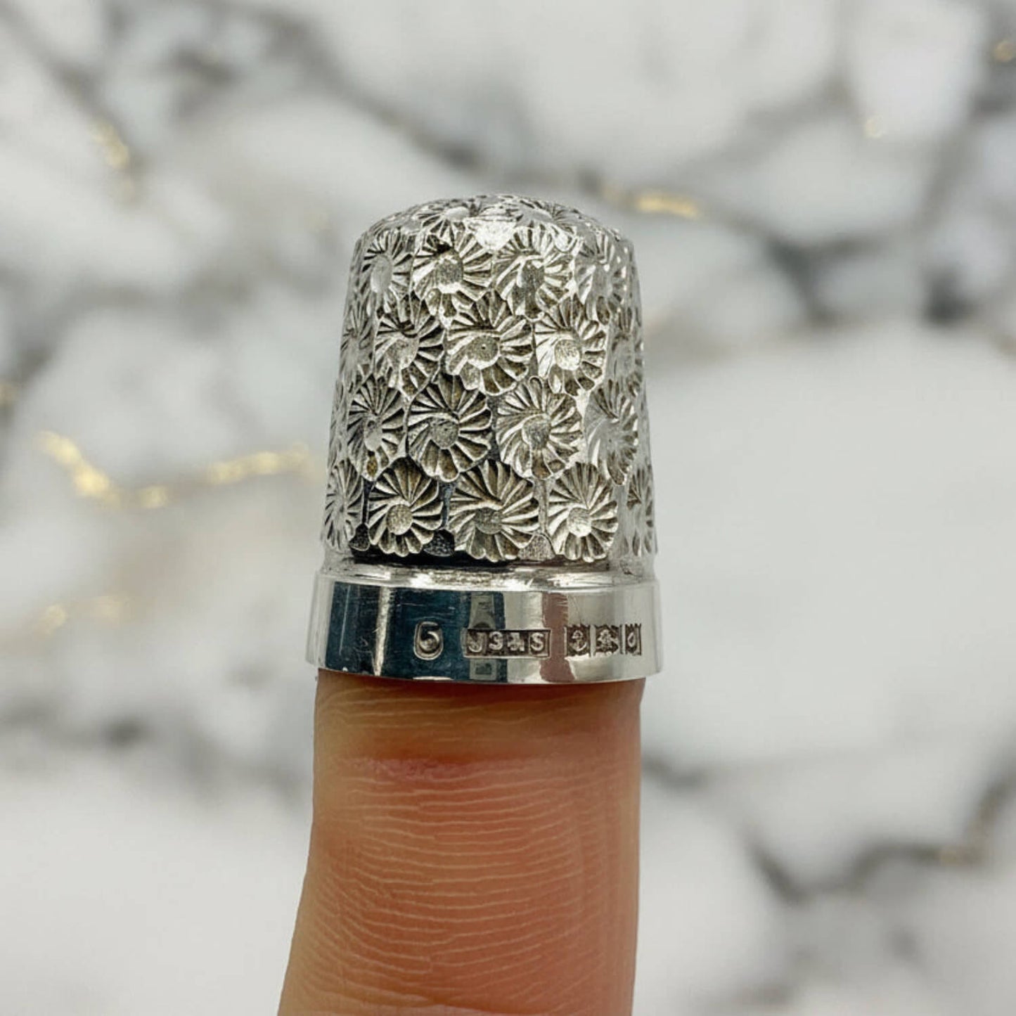 Silver thimble with floral pattern on a finger against a marble background