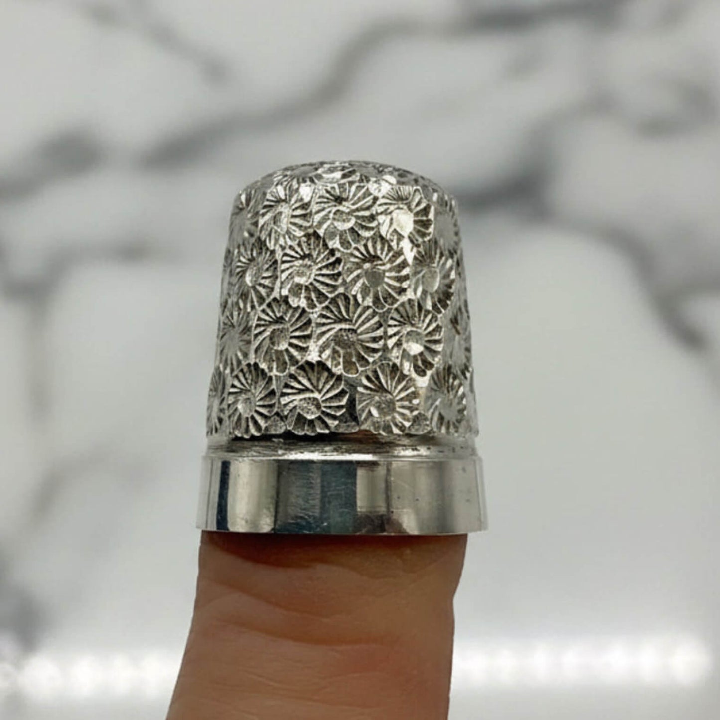 Silver thimble with floral pattern on a finger against a blurred background