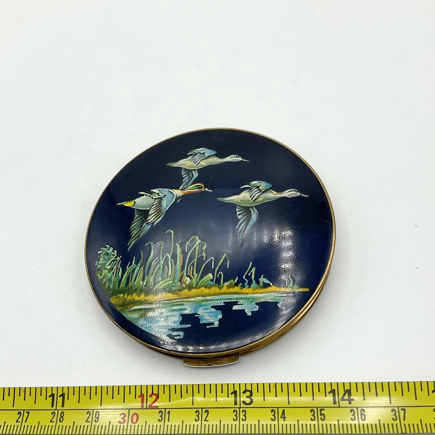 beautiful enamel featuring three ducks flying over water on a mirror compact lid next to a tape measure