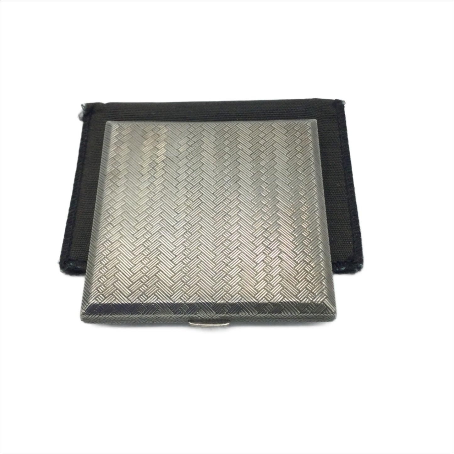A silver coloured square compact with a woven pattern to the lid. It is sitting on a black bag.