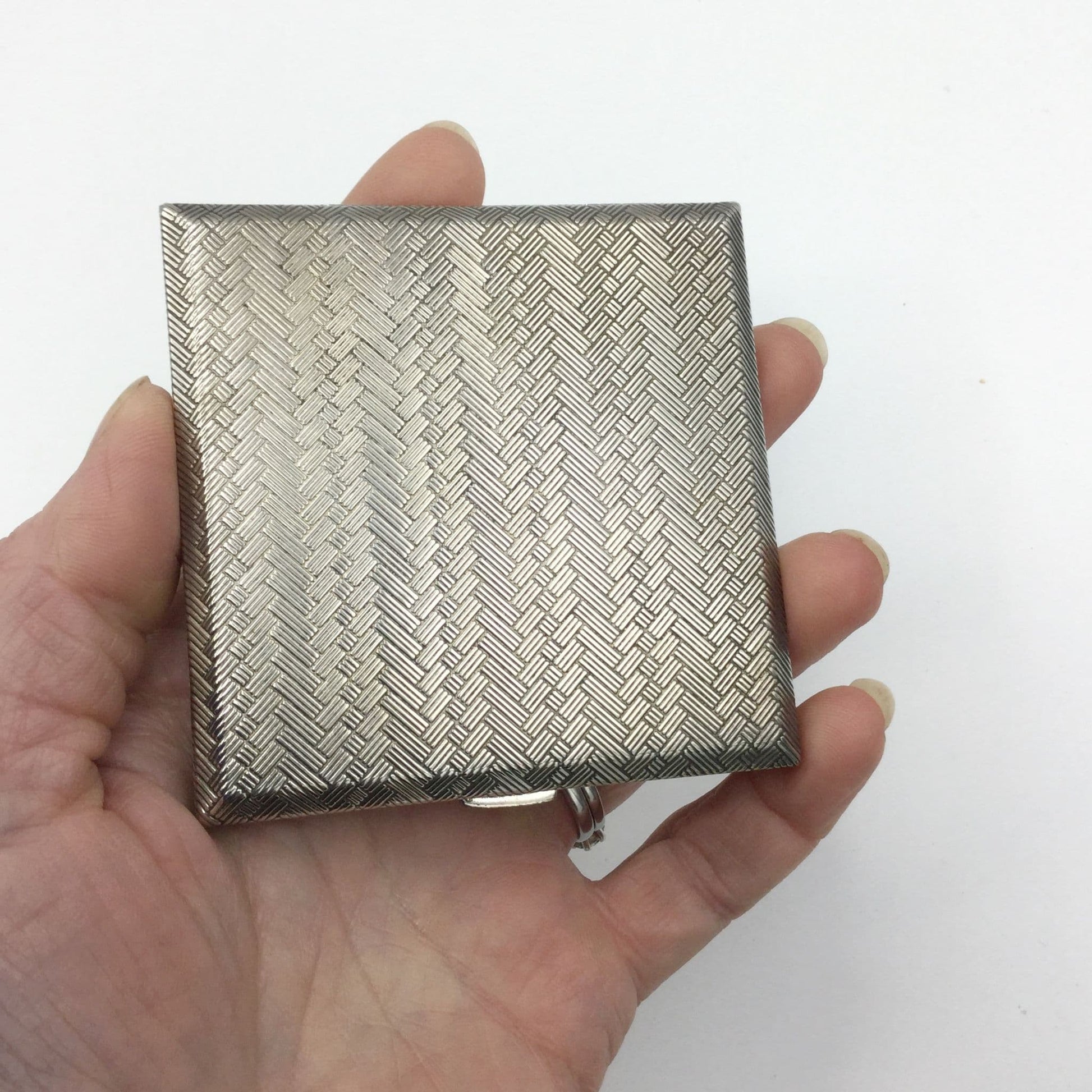 A silver coloured square compact with a woven pattern to the lid. It is being held in a hand.