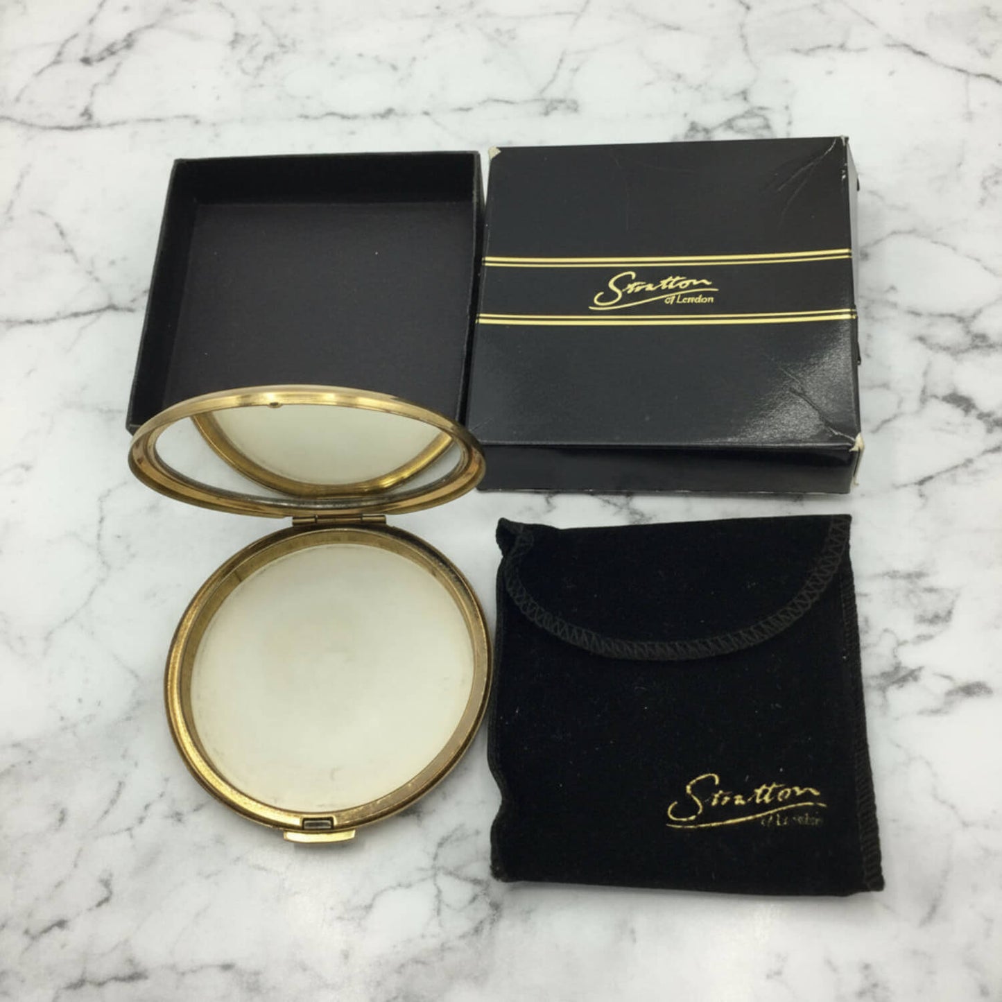 A round compact mirror with a gold-toned frame shown open, alongside its black presentation box and a matching black velvet pouch with gold lettering, all placed on a white marble surface.