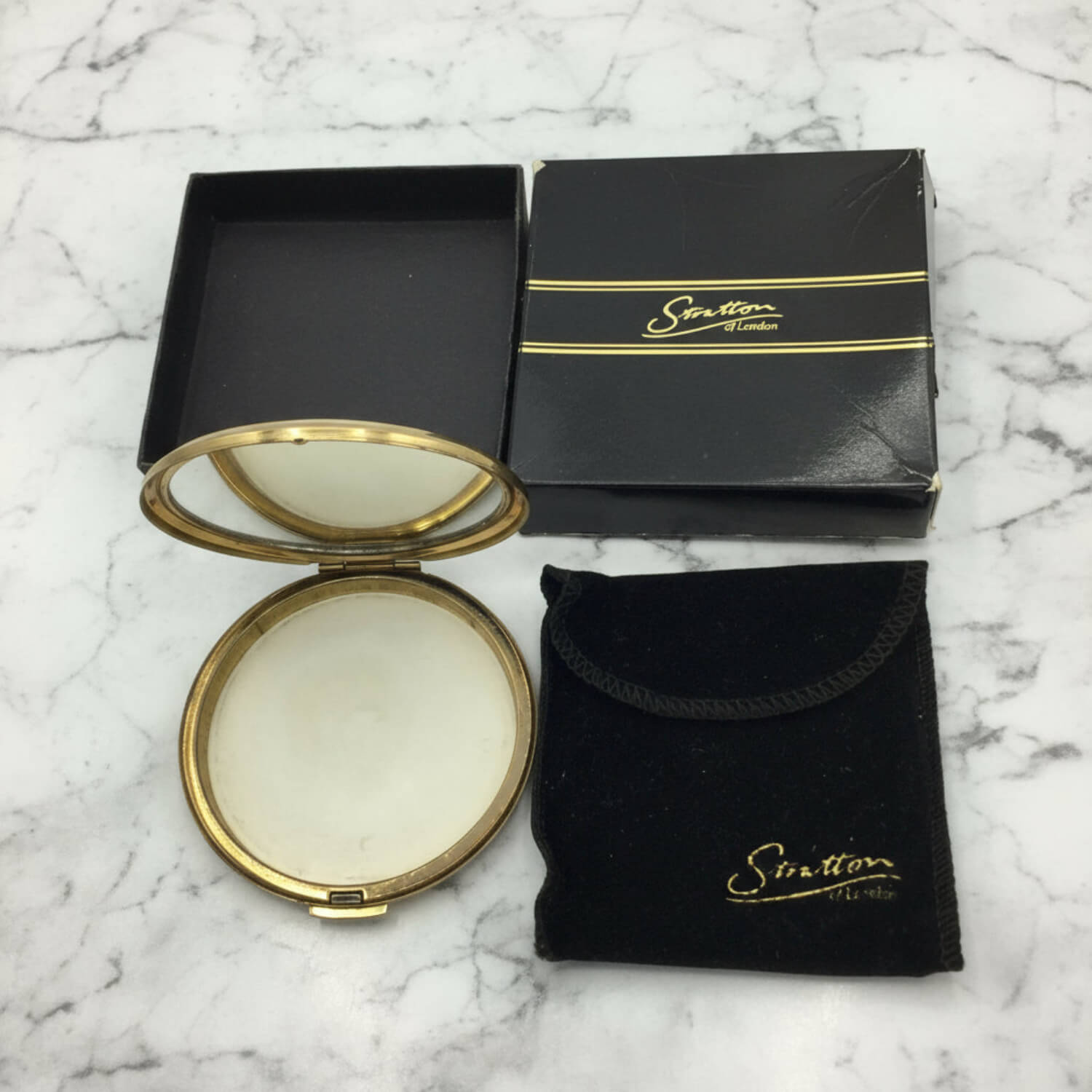 A round compact mirror with a gold-toned frame shown open, alongside its black presentation box and a matching black velvet pouch with gold lettering, all placed on a white marble surface.