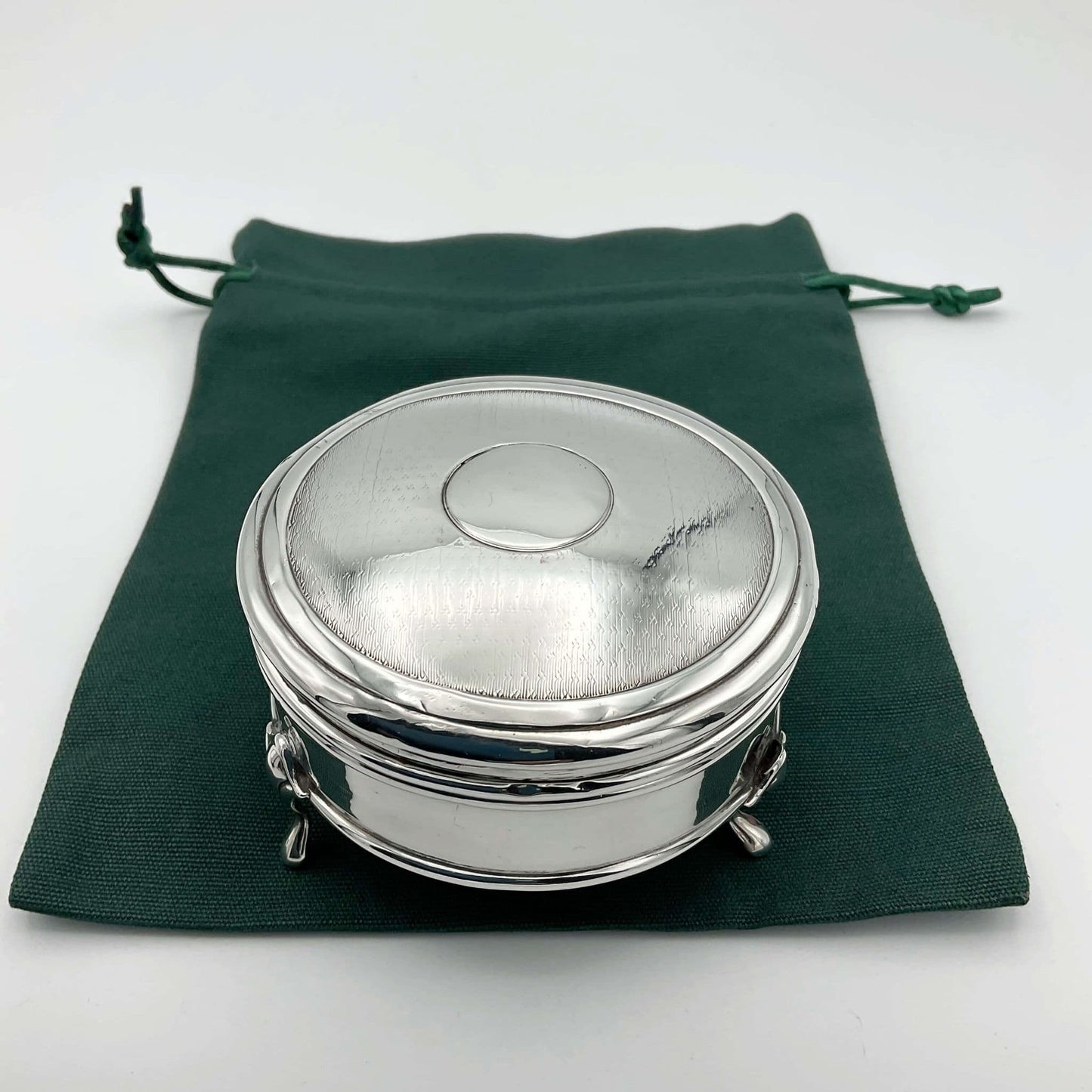 Round silver jewellery box with a pattern on the lid and legs standing on a green cotton bag.