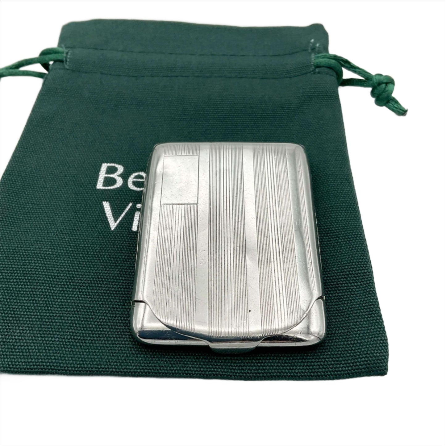 Silver matchbook case with a lined pattern and blank box for engraving initials in left hand corner. Case is on green Beeches Vintage cotton bag.