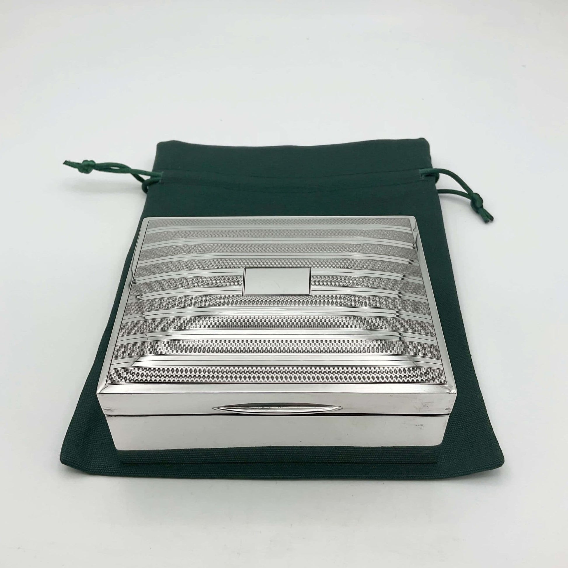 Vintage Sterling Silver Cigarette box with a striped pattern across the lid and a blank rectangular square in the centre. The cigarette  box is sitting on a green cotton bag.