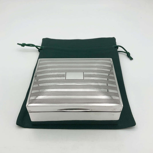 Silver box with an striped pattern across the lid and a blank rectangular square in the centre. The box is sitting on a green cotton bag.