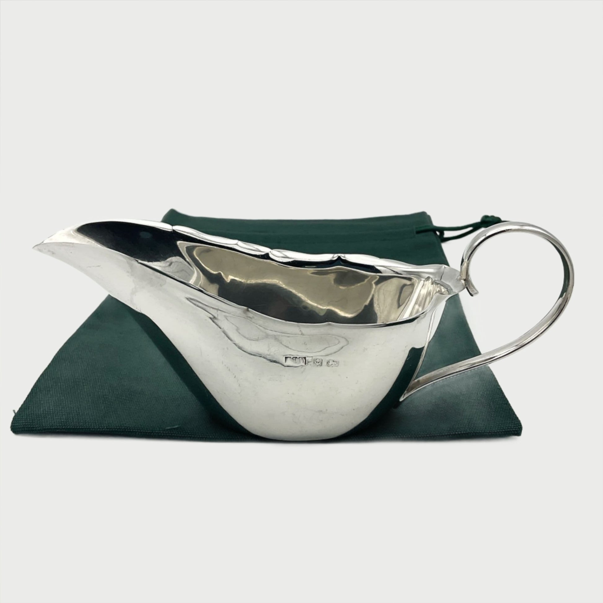 Shiny Silver sauce boat sitting on a green cotton bag. The sauce boat has a loop handle , crimped edges and Silver hallmarks on one side.