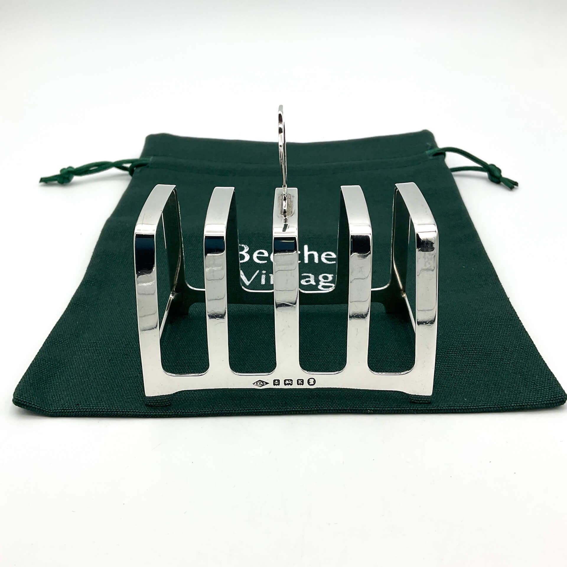 Art Deco Silver toast rack showing spaces for four slices of toast and a handle in the middle. The shiny toast rack is sitting on a green cotton bag.