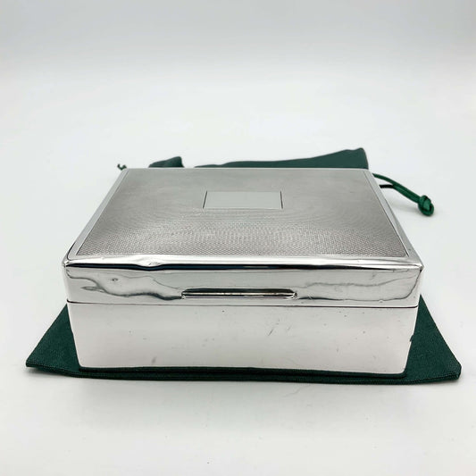 Silver box with shiny silver sides and a patterned lid sitting on a green cotton bag. The lid has a blank rectangular square in the centre. 