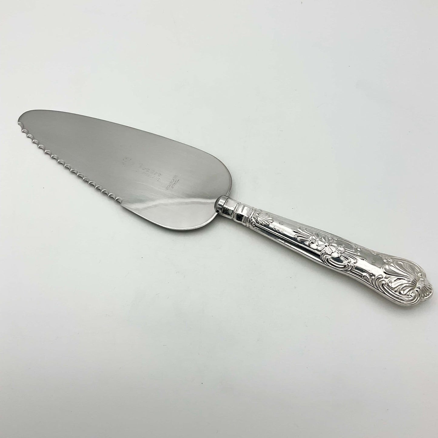 A silver coloured metal cake or pie slice on a white background. The handle has a Queens pattern elaborate design.