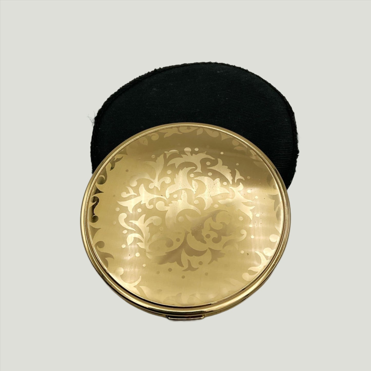 gold coloured lid of powder compact with leaf pattern on a black felt case