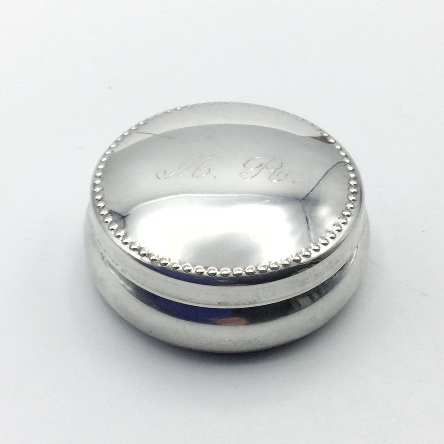 Round silver case with a dotted pattern around the lid and MR engraved in the centre on a white backgorund